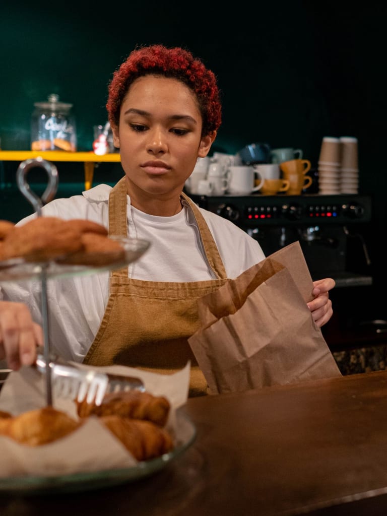 Młoda baristka z rudymi lokami, w lnianym fartuchu, pakuje szczypcami wypieki do papierowej torebki przy ladzie kawiarni