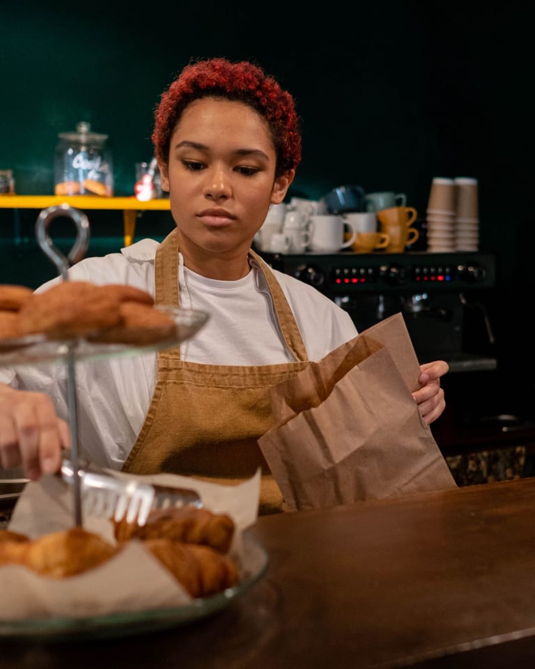 Młoda baristka z rudymi lokami, w lnianym fartuchu, pakuje szczypcami wypieki do papierowej torebki przy ladzie kawiarni