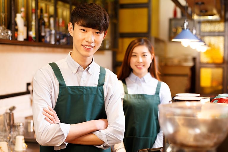 Zwei lächelnde Servicekräfte mit grünen Schürzen stehen hinter der Theke eines Cafés