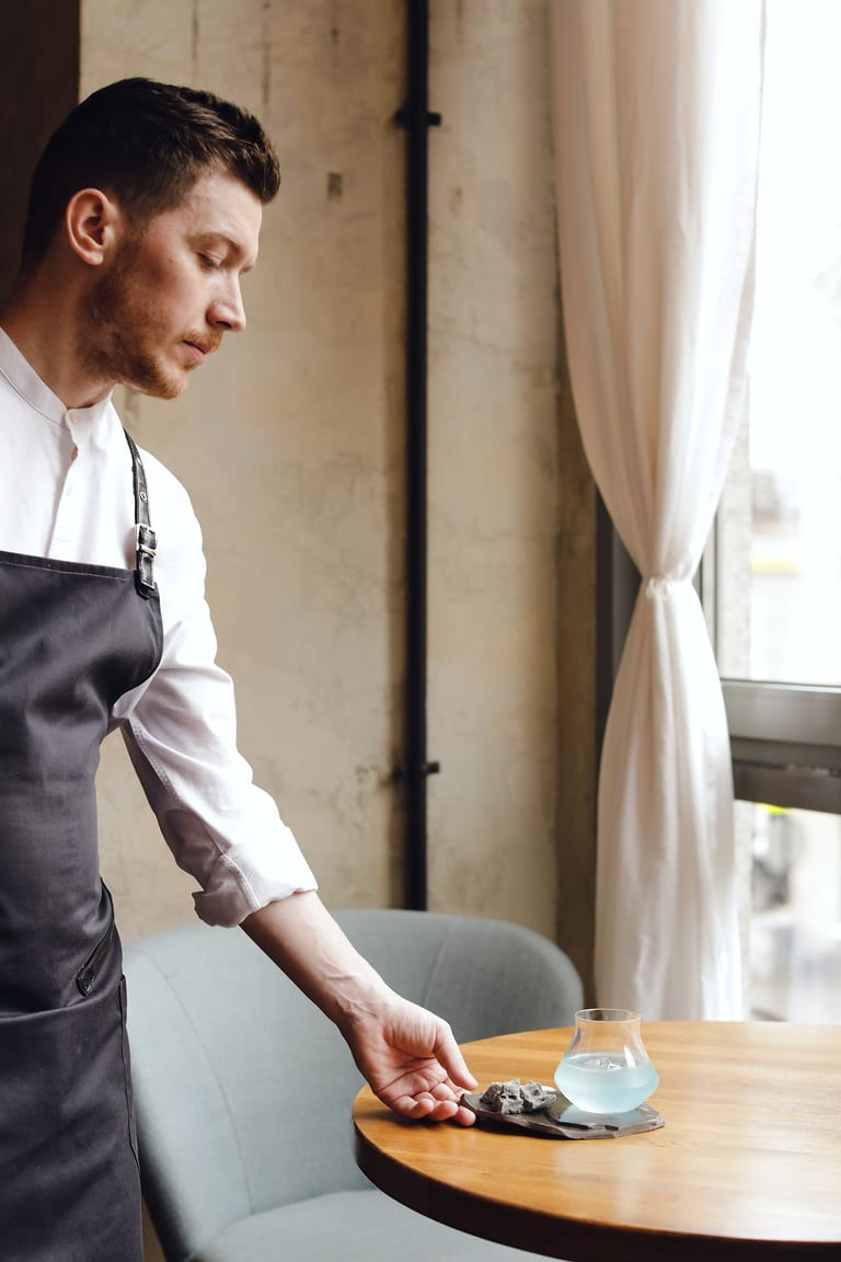 Kellner mit dunkler Schürze serviert in einem Lokal am Fenster ein Getränk auf einem Schieferbrett an einem runden Holztisch