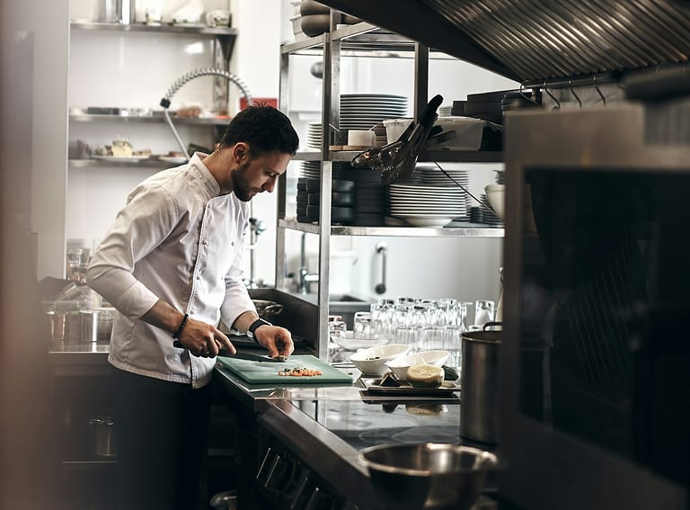 Junger Koch in weißer Kochjacke schneidet in einer Restaurantküche Gemüse auf einem grünen Schneidebrett