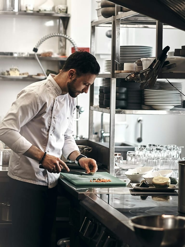 Młody kucharz w białej bluzie kucharskiej kroi warzywa na zielonej desce w kuchni restauracji