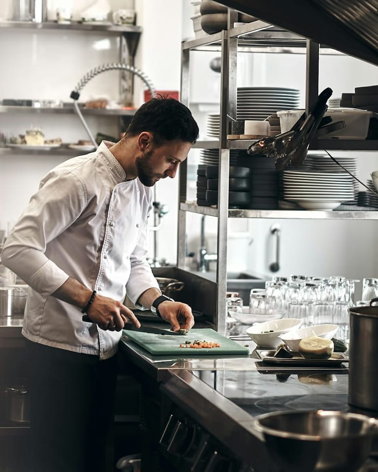 Młody kucharz w białej bluzie kucharskiej kroi warzywa na zielonej desce w kuchni restauracji