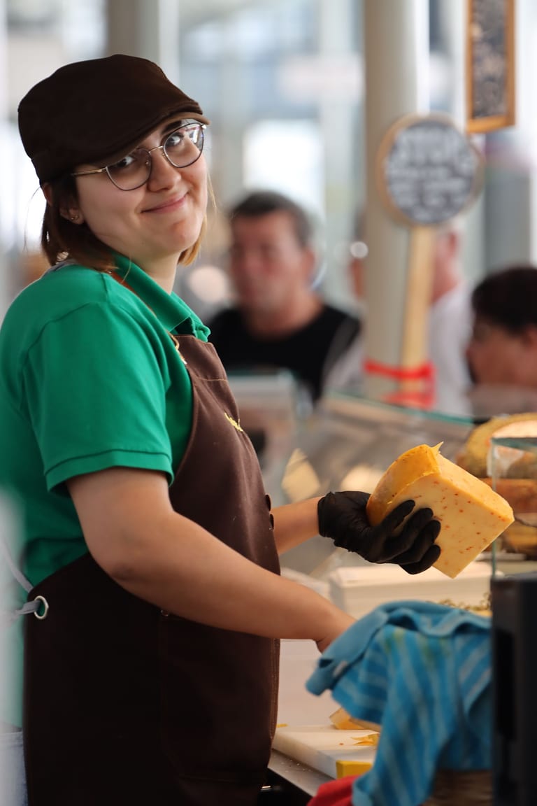 Junge Verkäuferin mit Brille, Kappe und brauner Schürze hält an der Käsetheke einer Markthalle ein Stück Käse