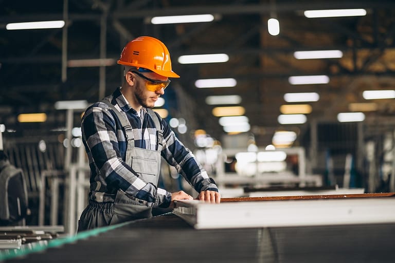 Arbeiter mit orangem Schutzhelm und Schutzbrille arbeitet an einem Werktisch in einer Produktionshalle