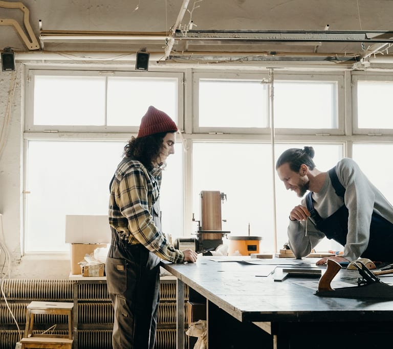Zwei junge Männer arbeiten an einer Werkbank in einer hellen Holzwerkstatt mit großen Sprossenfenstern