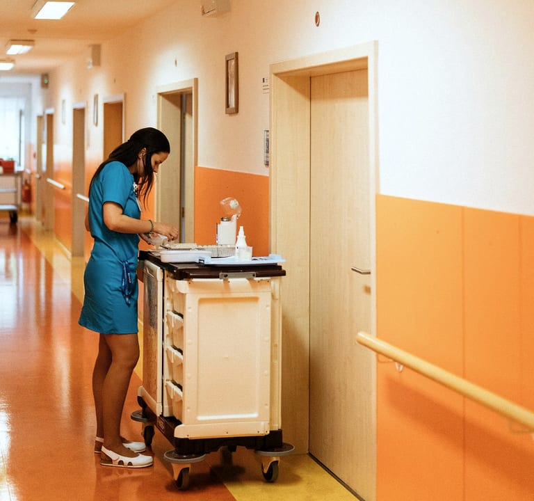 Junge Pflegerin in türkisfarbener Dienstkleidung bereitet im Flur einer Pflegeeinrichtung am Medikamentenwagen Medikamente vor