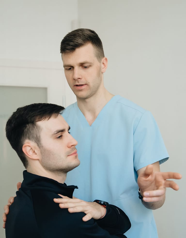 Junger Physiotherapeut in hellblauer Dienstkleidung korrigiert in einer Therapiepraxis die Schulterhaltung eines jungen Patienten