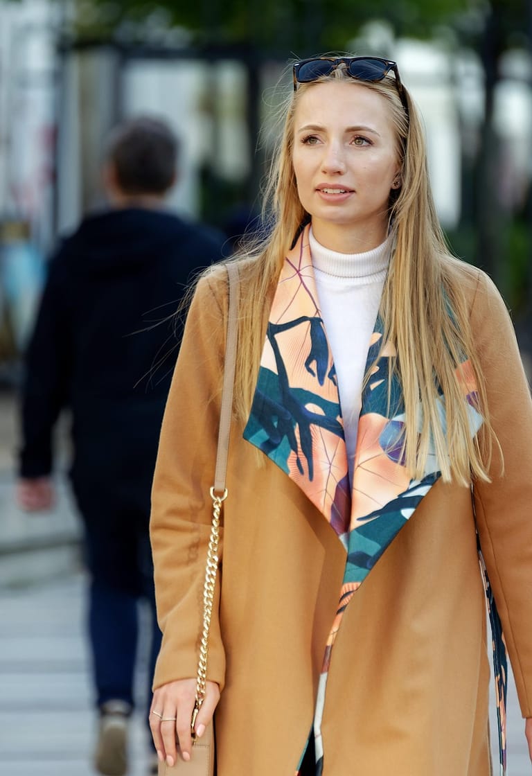Junge Frau mit langen blonden Haaren, Sonnenbrille im Haar und camelfarbenem Mantel geht eine Straße entlang