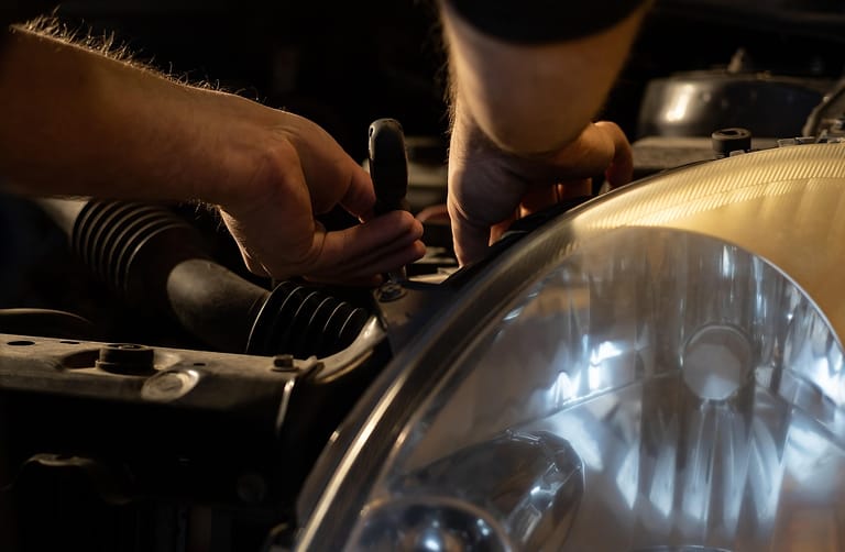 Hände arbeiten mit einem Werkzeug im Motorraum eines Autos neben einem Scheinwerfer, warmes Licht auf dunklem Grund