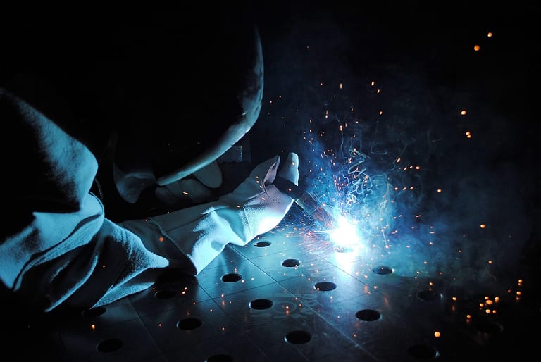 Schweißer mit Schutzhandschuh arbeitet an einer gelochten Metallplatte, blaues Licht und Funken vor dunklem Hintergrund