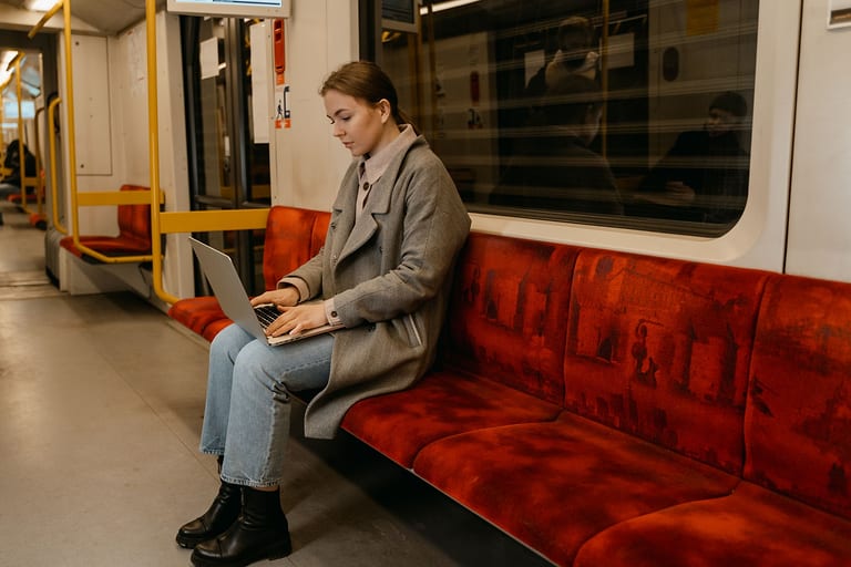 Junge Frau im grauen Mantel arbeitet mit dem Laptop auf dem Schoß in einem U-Bahn-Wagen