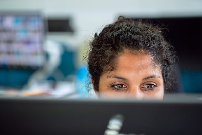 Frau mit lockigem dunklem Haar blickt konzentriert über einen Bildschirm in einem Büro