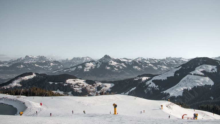 Skipiste mit Schneekanonen und einzelnen Skifahrern im Skigebiet bei Brixen im Thale, dahinter verschneite Berge
