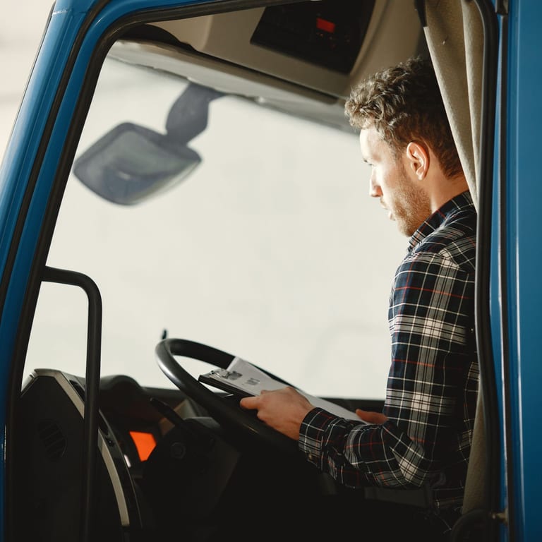 Junger Lkw-Fahrer im karierten Hemd sitzt in der Fahrerkabine eines blauen Lkw und prüft Frachtpapiere auf dem Klemmbrett